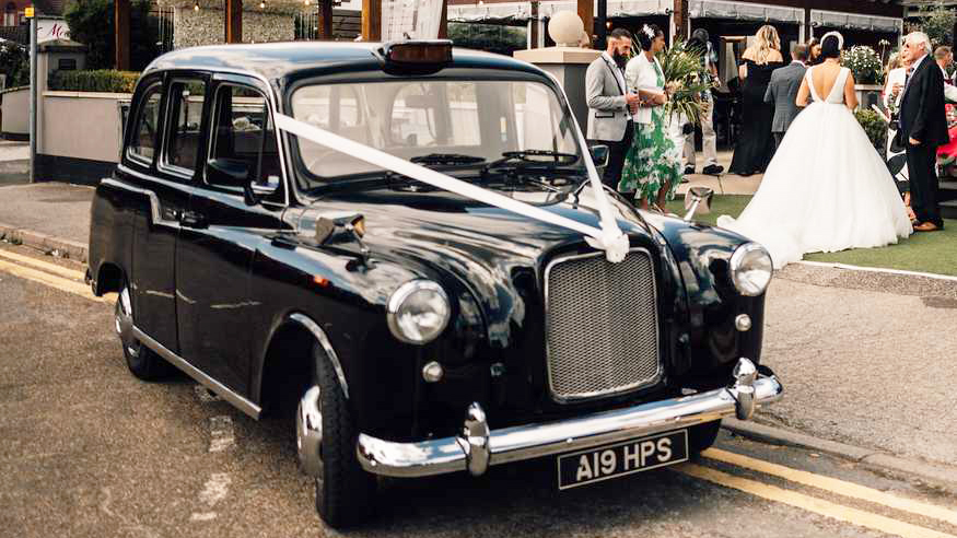 Black classic taxi cab decorated with white ribbons. Bride and wedding guests in the background