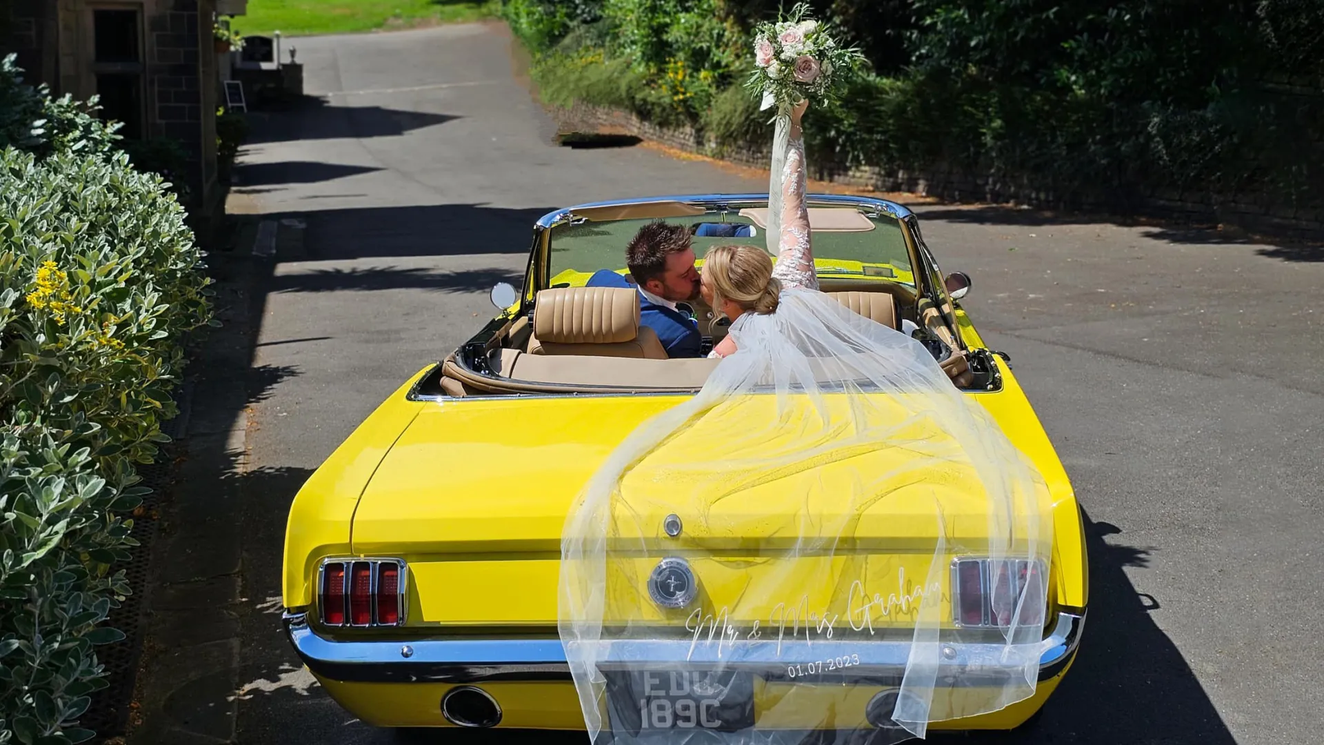 Bride and Groom seating inside a convertible yellow Mustang with Bride holding her bouquet of flowers high in the air