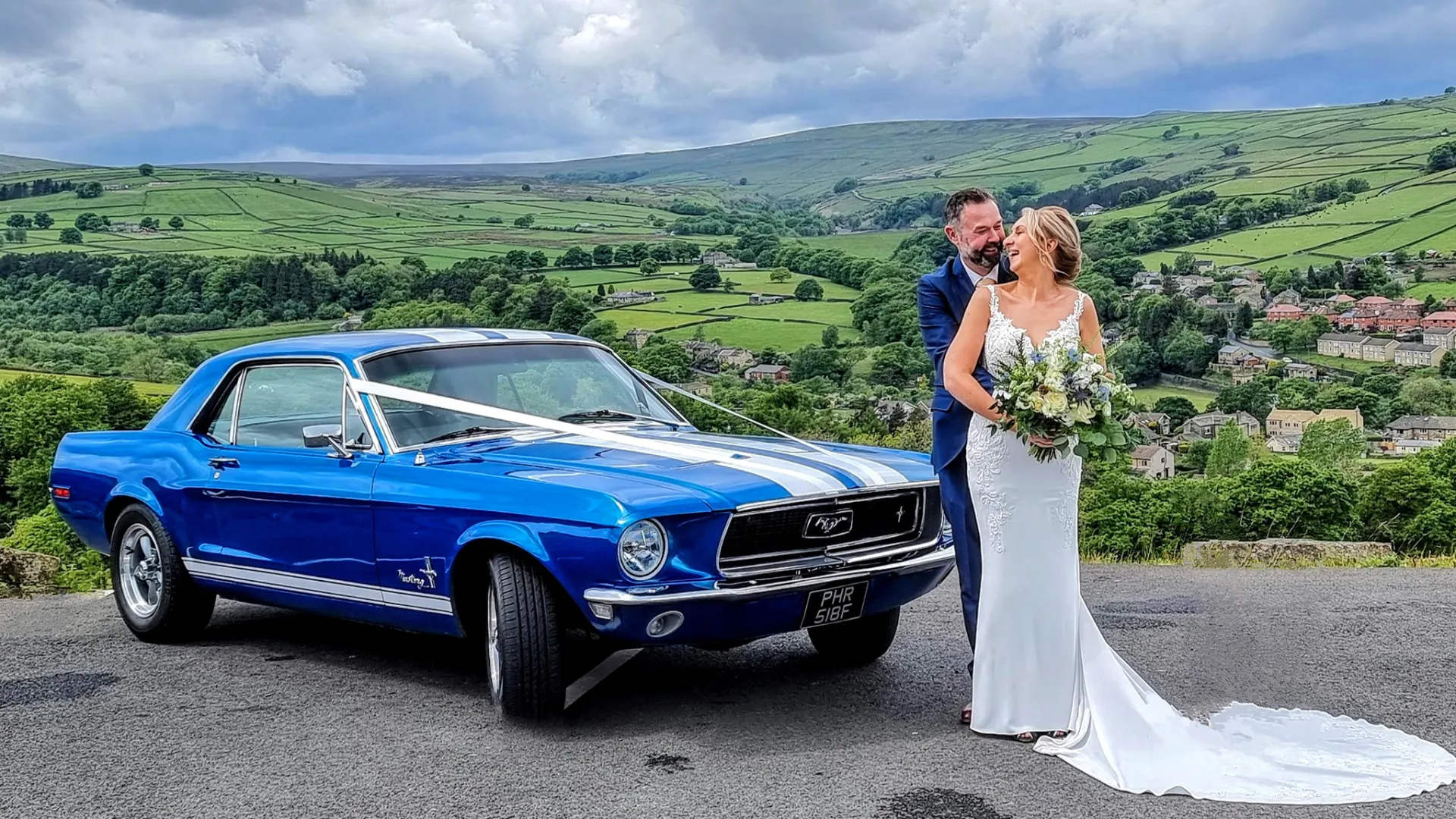 Classic Blue Ford Mustang dressed with White Ribbons across its bonnet. Bride and Groom standing in front of the car holding each others.