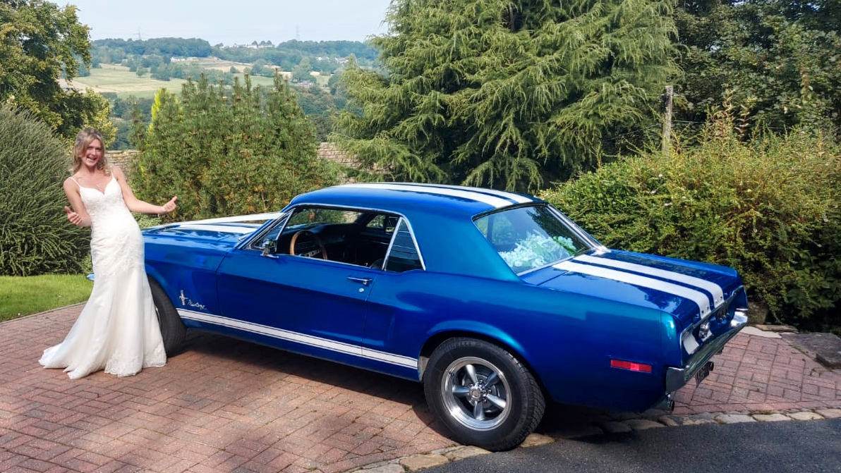 Bride wearing a white dress standing by a Classic Blue Ford Mustang