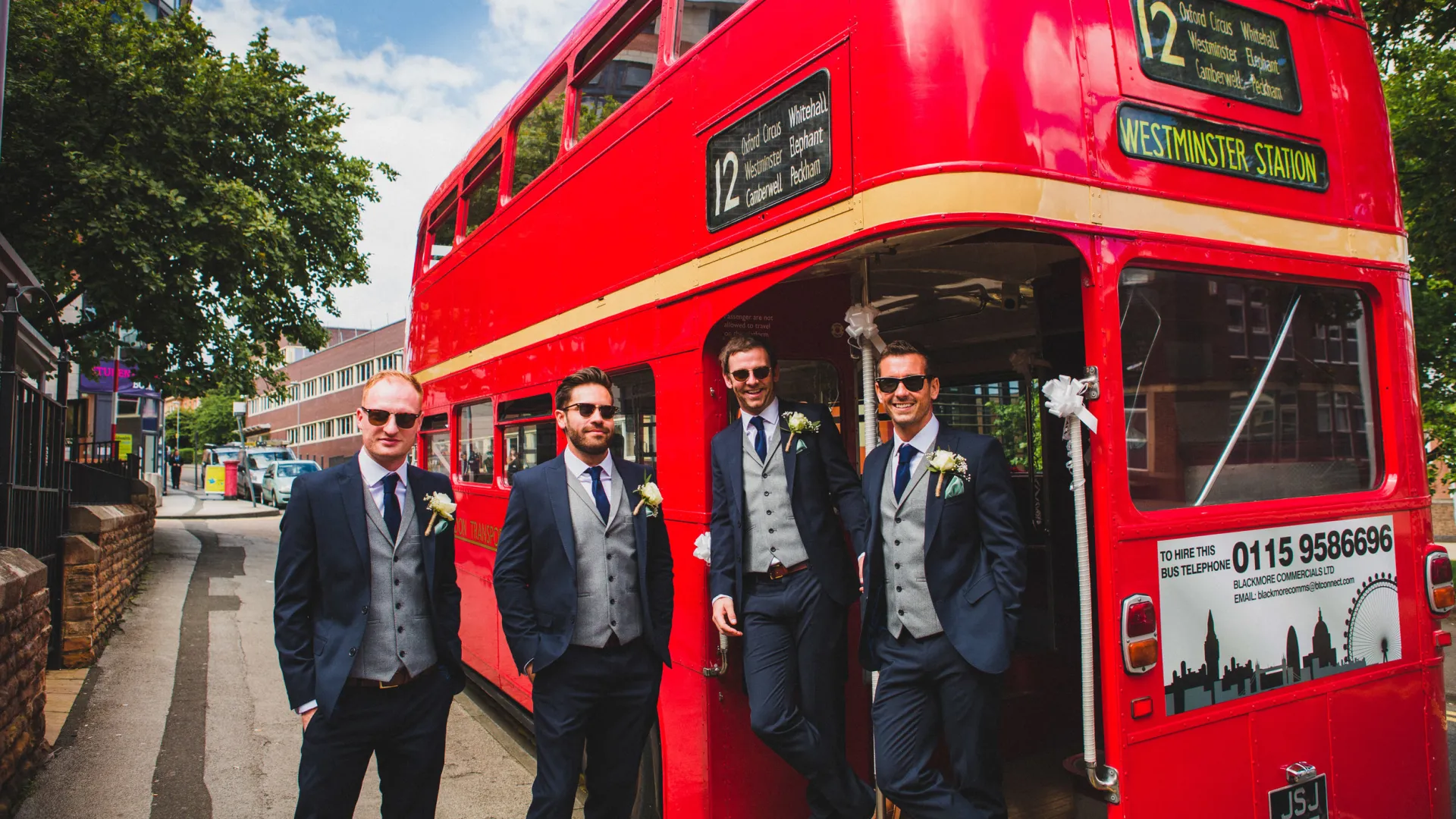 Groom and his three groomsmen standing on the rear open platform of a Classic Double Decker Red Bus