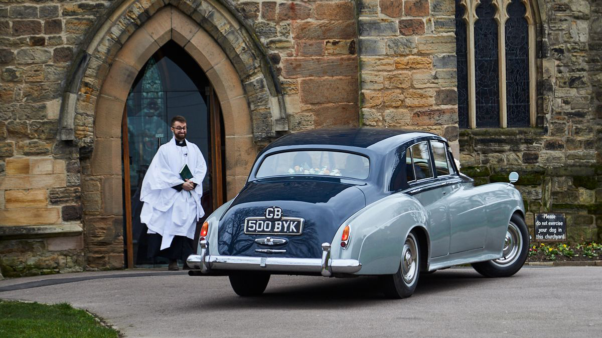 rear view of a Classic Black & Silver Rolls-Royce silver Cloud in front of chruch with priest in the background.