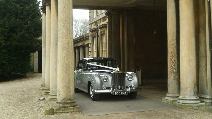 Classic Black & Silver Rolls-Royce silver Cloud with white ribbons in front of wedding venue's main entrance