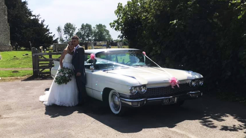 Bride and Groom standing by a Classic Cadilalc decorated with white and pink wedding ribbons