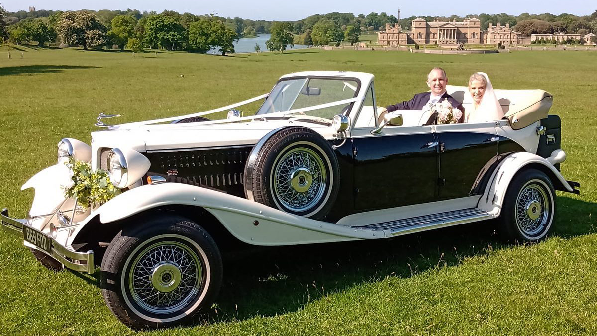 vintage Beauford Convertible in Black and White with Bride and Groom seating in the vehicle with Holkham Hall in Norfolk shown in the background