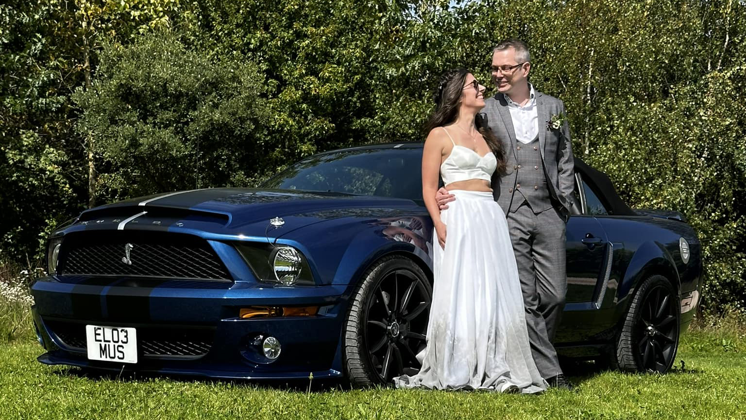 Bride and Groom standing in front of a dark blue modern Ford Mustang with green trees in the background