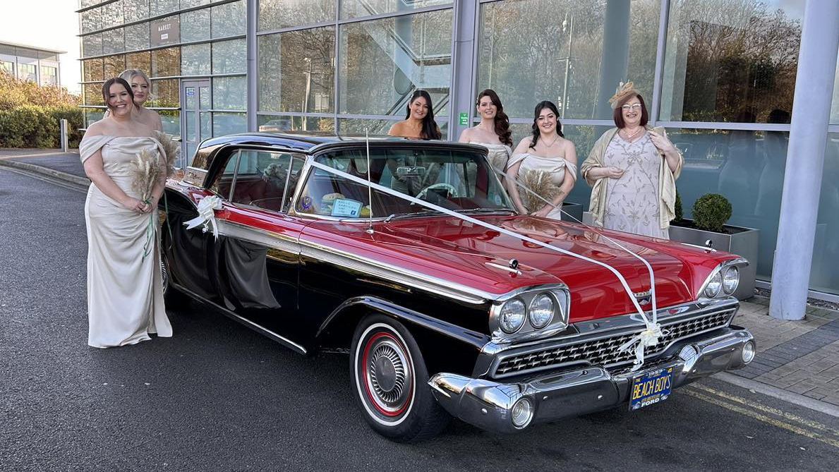 Bride and Bridesmaids standing by a classic american Ford Fairlane in Black and Red decorated with white wedding ribbons