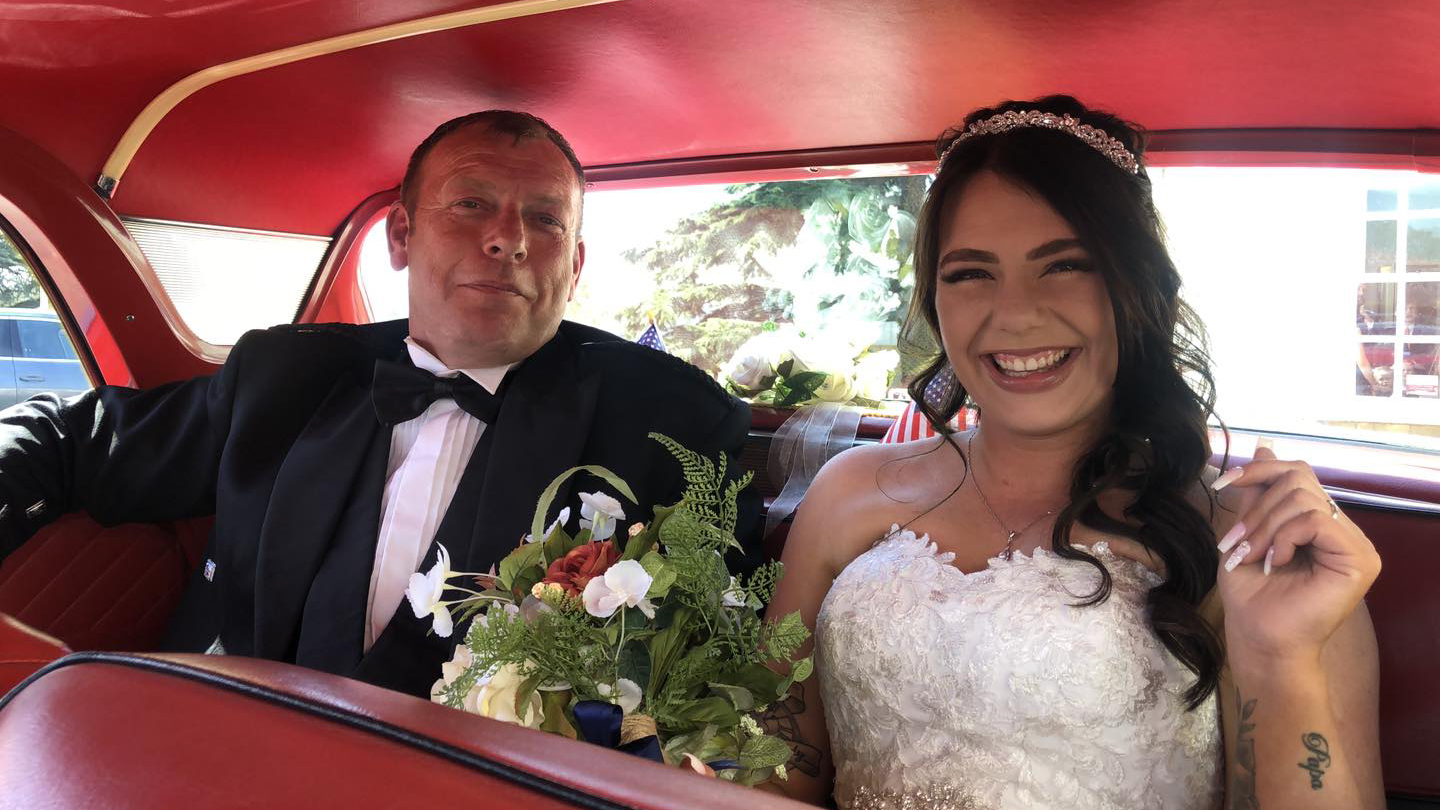 Bride and Father seating inside American Ford Fairlane. Smiling Bride is wearing a white dress and holding a bouquet of flowers.