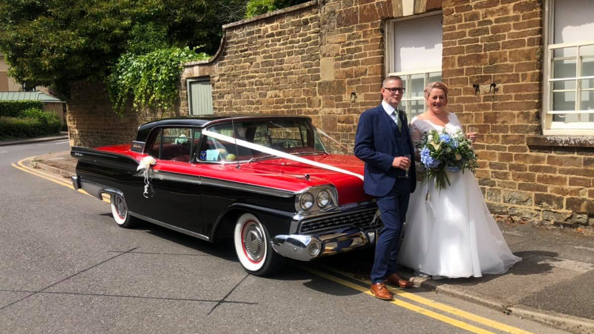 Bride and Groom standing in front of a classic american Ford Fairlane with white wedding ribbons accros its bonnet and white bows on door handle