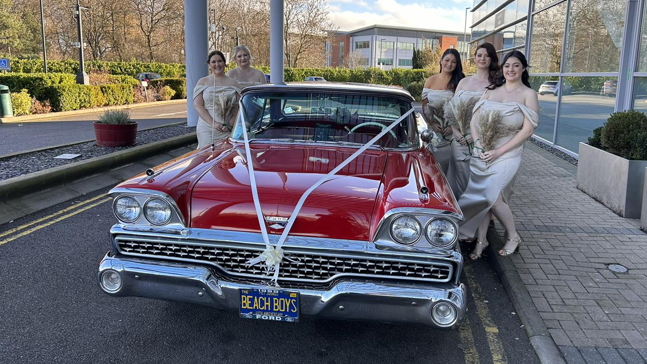 American Ford Fairlane in Black and Red with white wedding ribbons in front of wedding venue with Bride and Bridesmaids standing by the cars in matching Ivory dresses