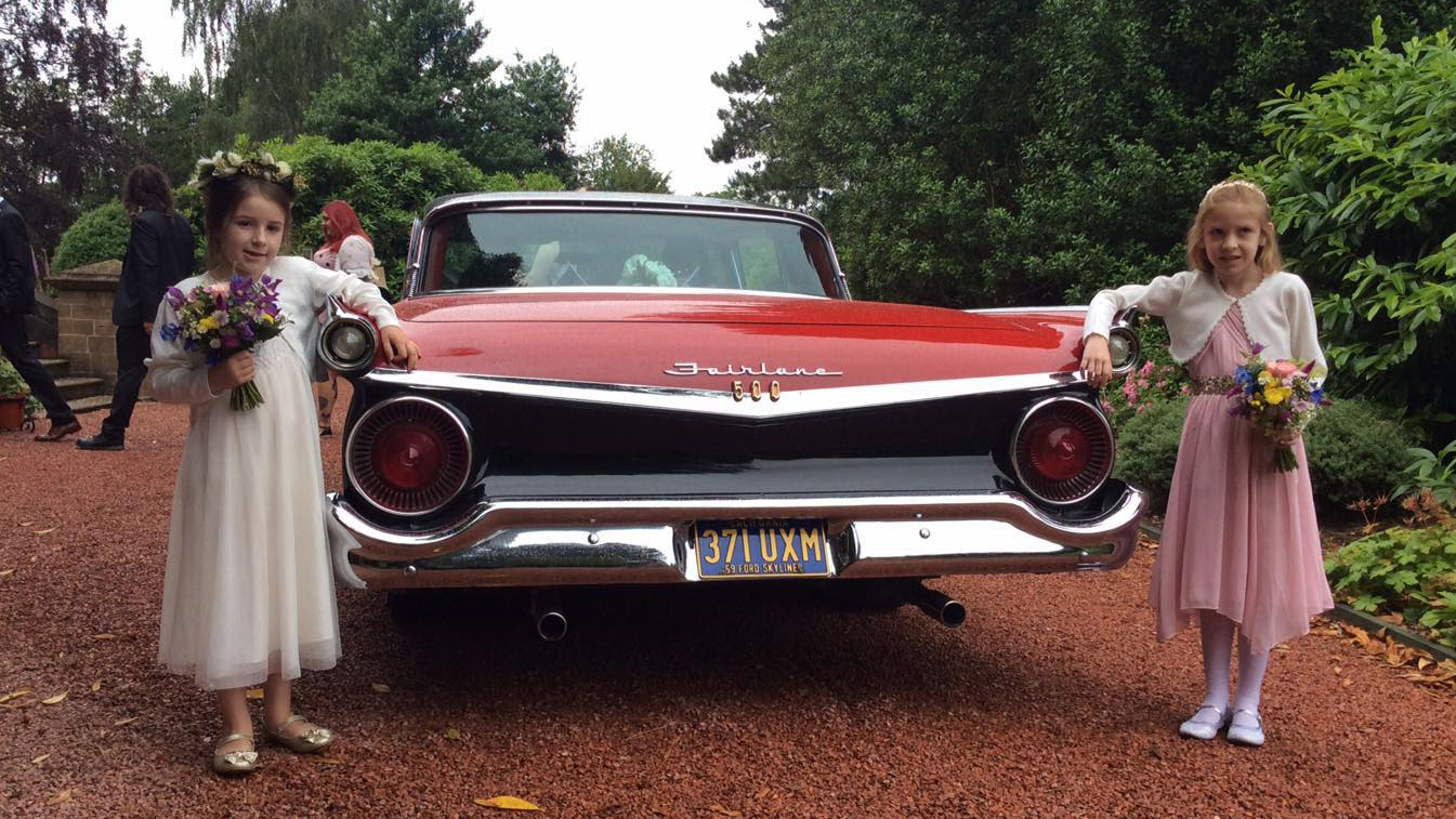 Two young flower girls standing on both sides of a classic Ford Fairline