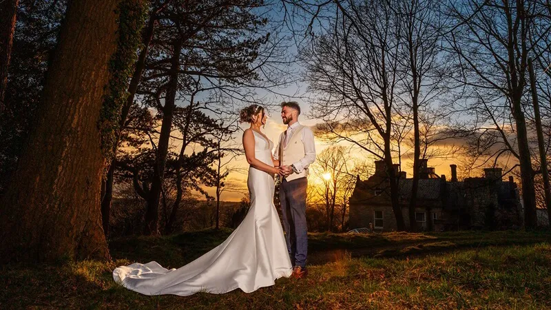 Bride wearing a white dress holding hands with her groom in an Autumn coloured forest edge. Hollin House located in Cheshire can be seen in the background and a stunning Twilight sunset in the background.