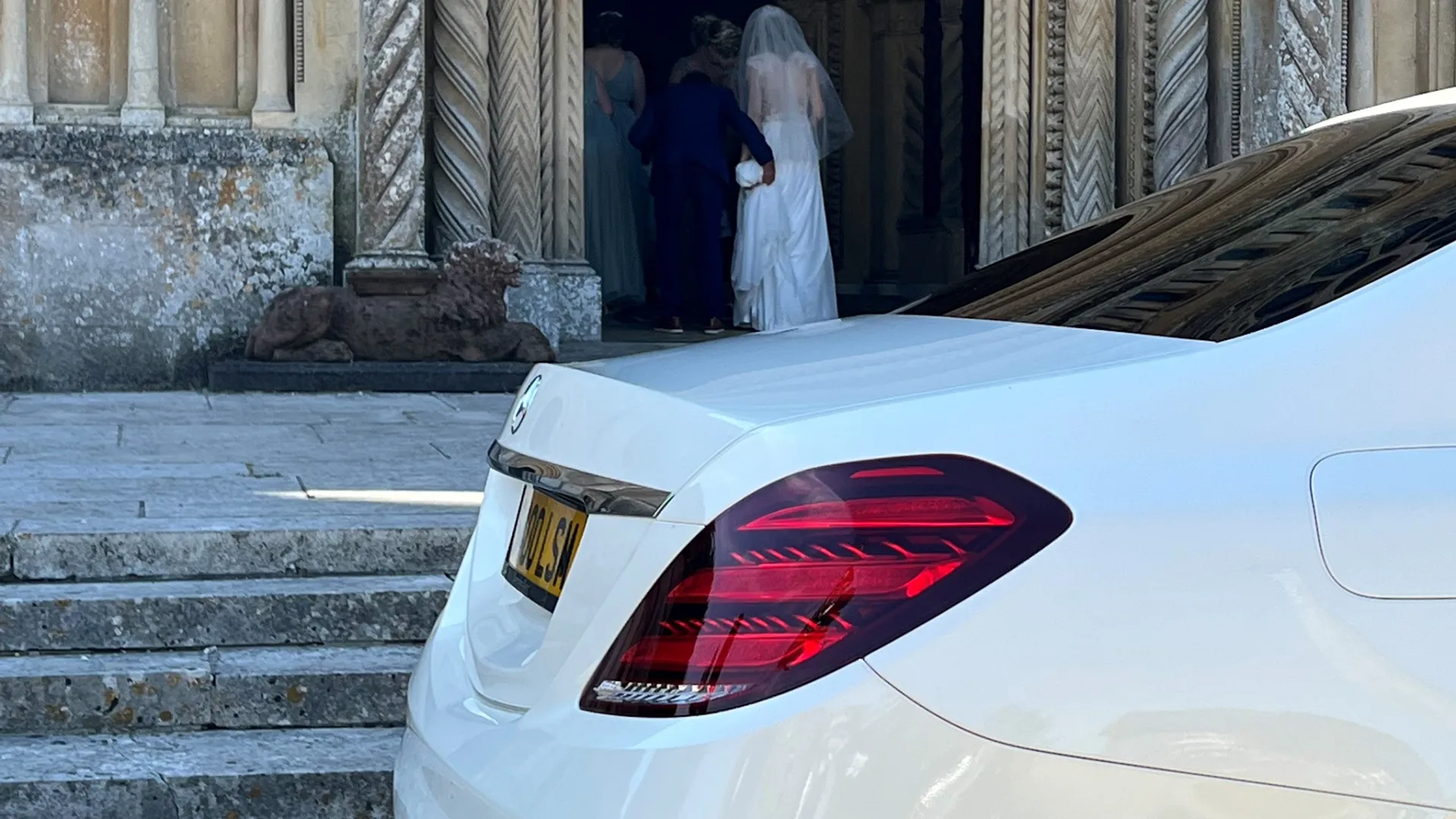 Rear part of a white mercedes in the foreground and Bride waering a white wedding dress walking into a Church in the background.