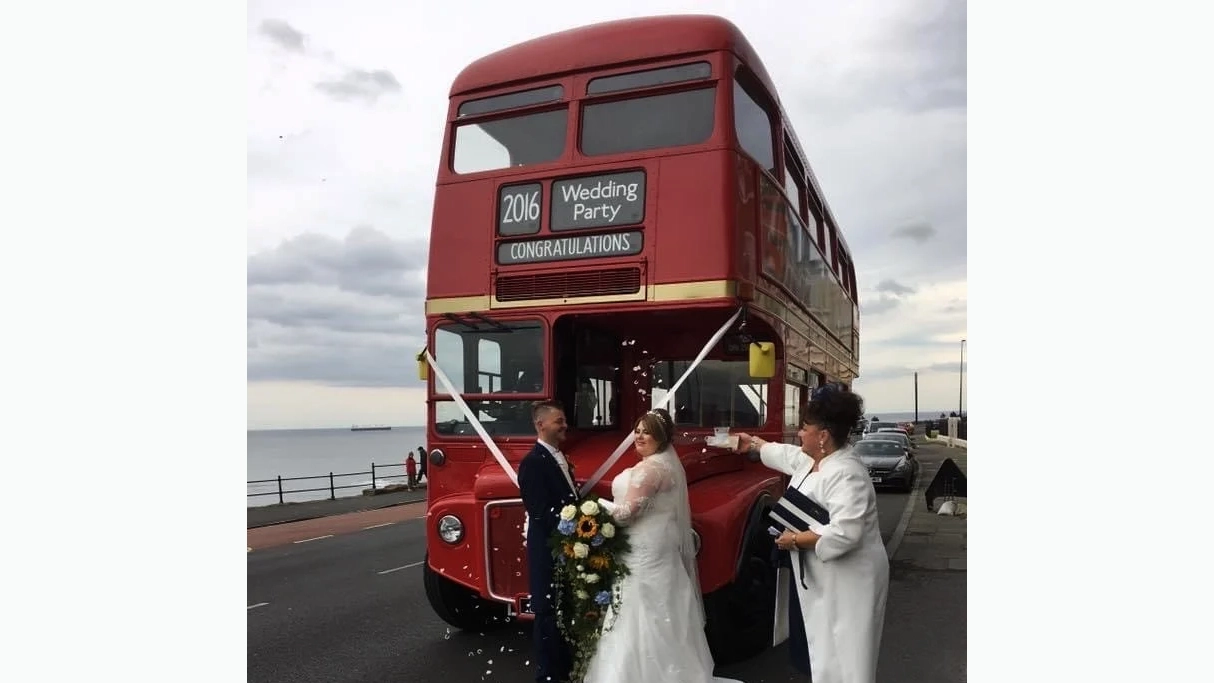 Bride and Groom standing in front of a Bid red routemaster bus with wedding guests throwing confettis