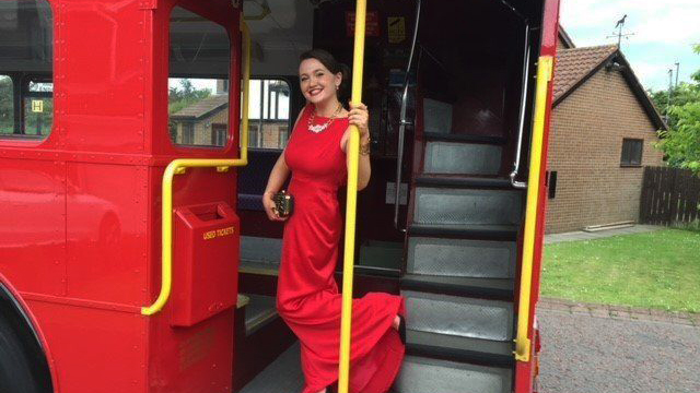 A smiling Wedding Guests wearing a red dress standing on the rear open platform of a Routemaster Bus