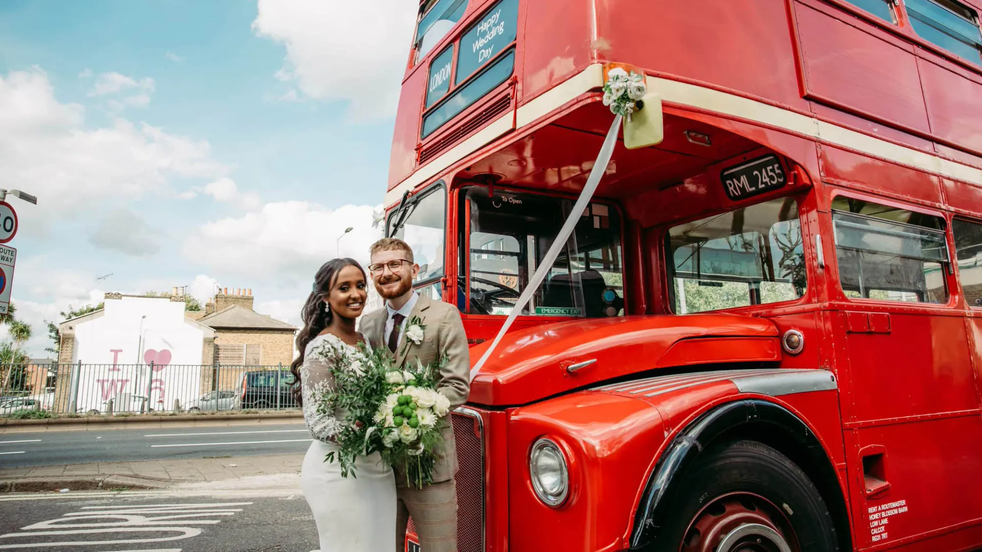 Bride and Groom standing in front of an old Red London Routemaster bus deocrated with Ribbons. Bride is holding her wedding bouquet in her hands