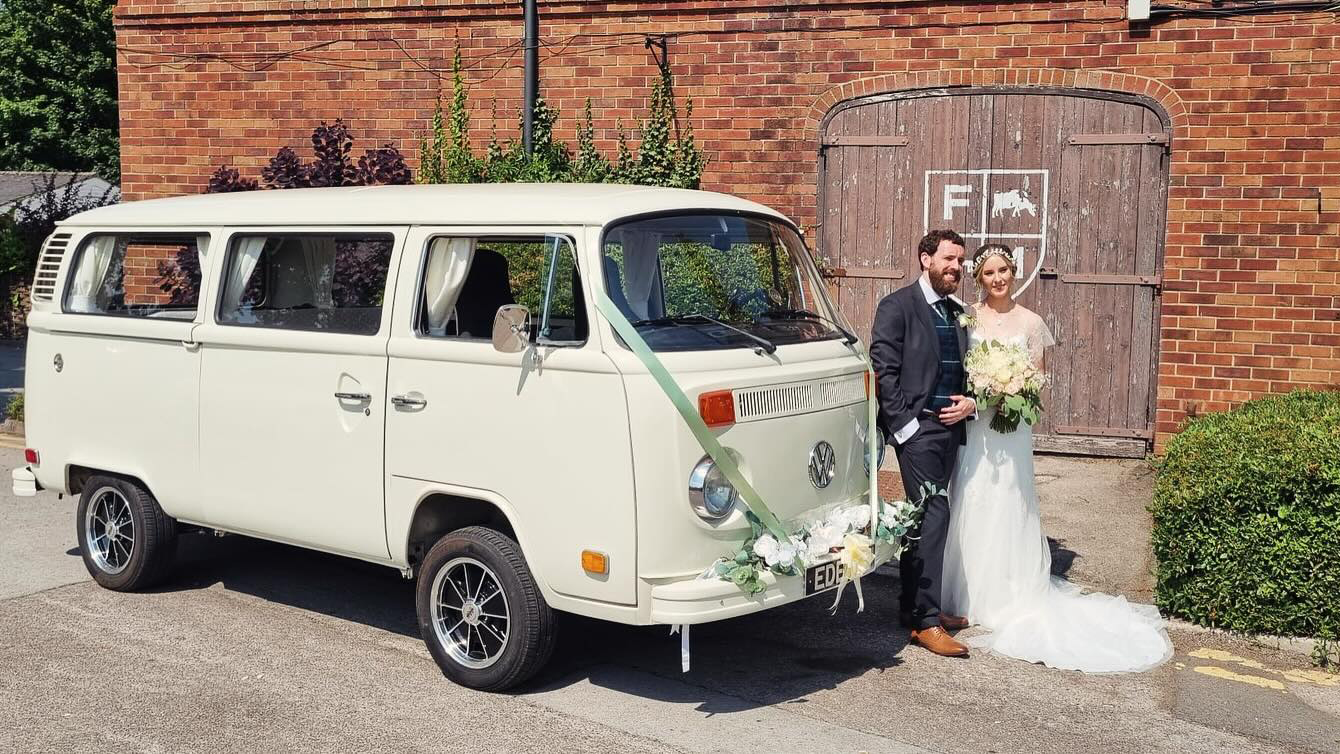 Classic VW campervan decorated with wedding ribbons with Bride and Groom standing in front of the vehicle