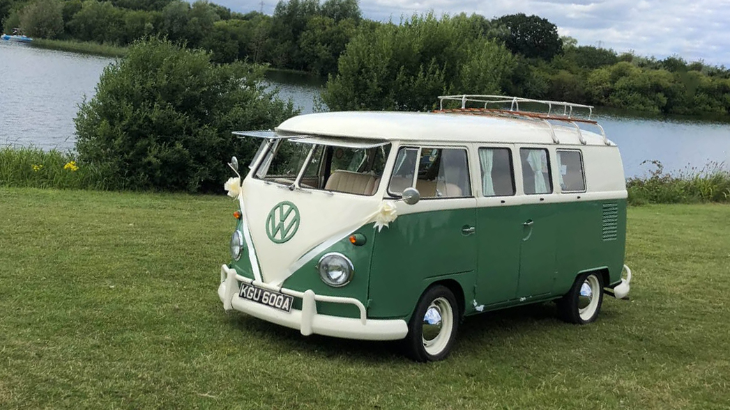 Retro Classic VW splitscreen campervan in Green and White with its front windows open parked in a green park with a lac in the background