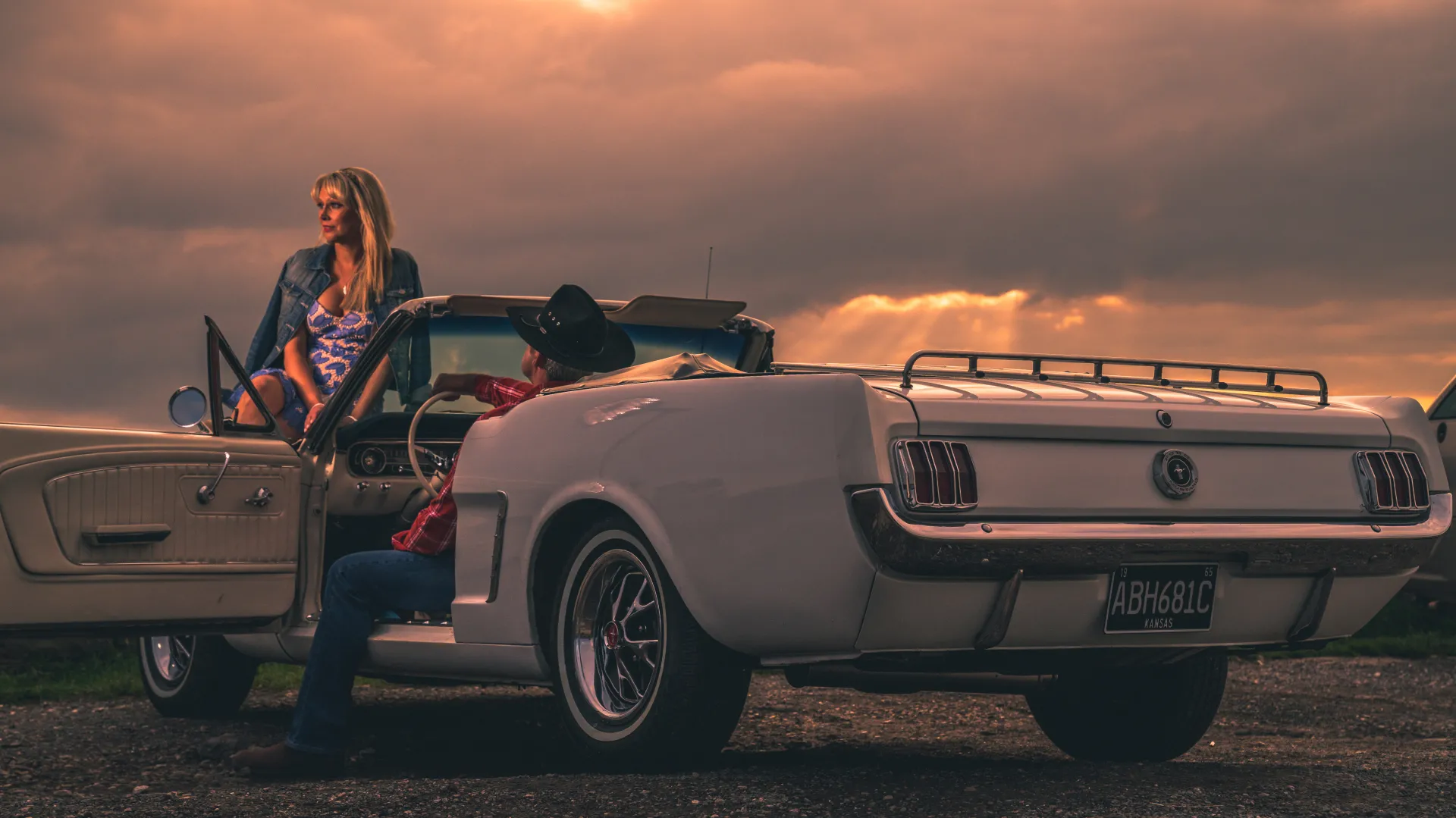 Rear view of the classic Ford Mustang at dusk with a man seating in the driver's seat with a cowboy hat and woman seating on the front hood.