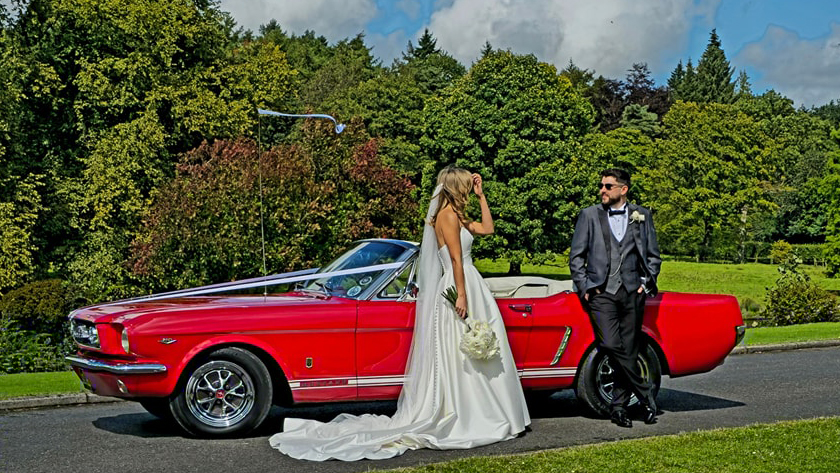 Bride wearing a white dress standing by a classic Ford Mustang Convertible in Red with her groom