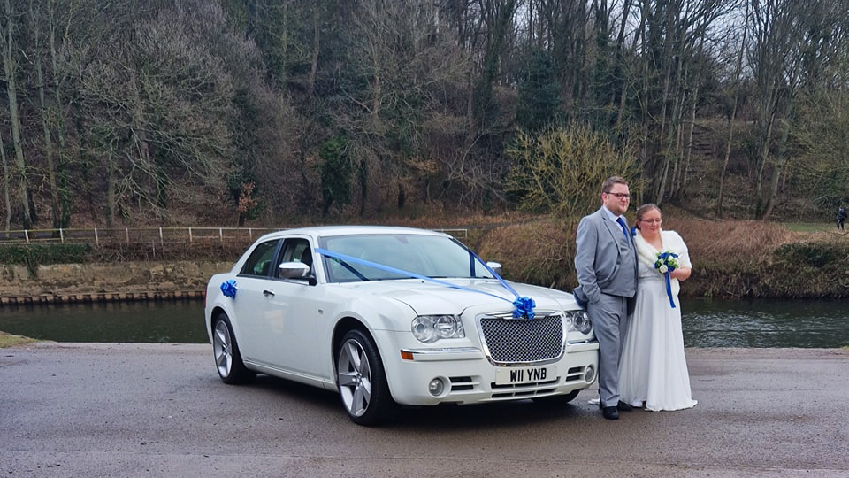 Bride and Groom standing by a White Chrysler 300c Saloon Car decorated with Blue Ribbons and bows