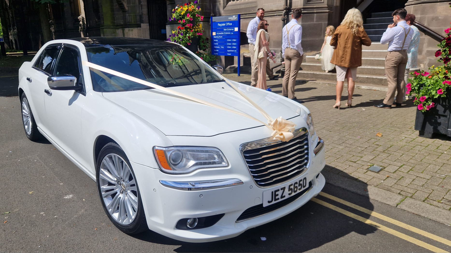 White chrysler 300c saloon car in front of a wedding church and dressed with light organe ribbons