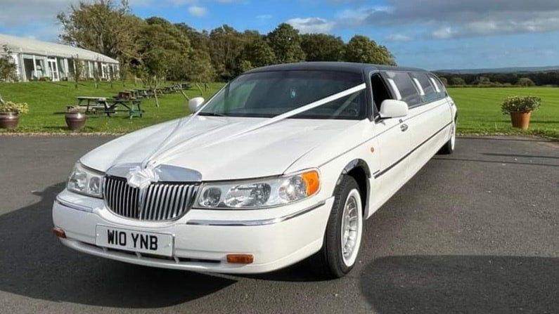 White Stretched Lincoln Limousine with black Roof decorated with pale pink wedding ribbons parked at a wedding venue