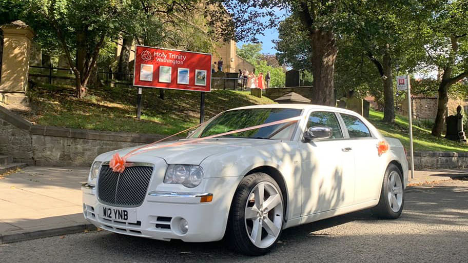 White Chrysler 300c Saloon Car in front of a church in Durham decorated with light orange ribbons and bows