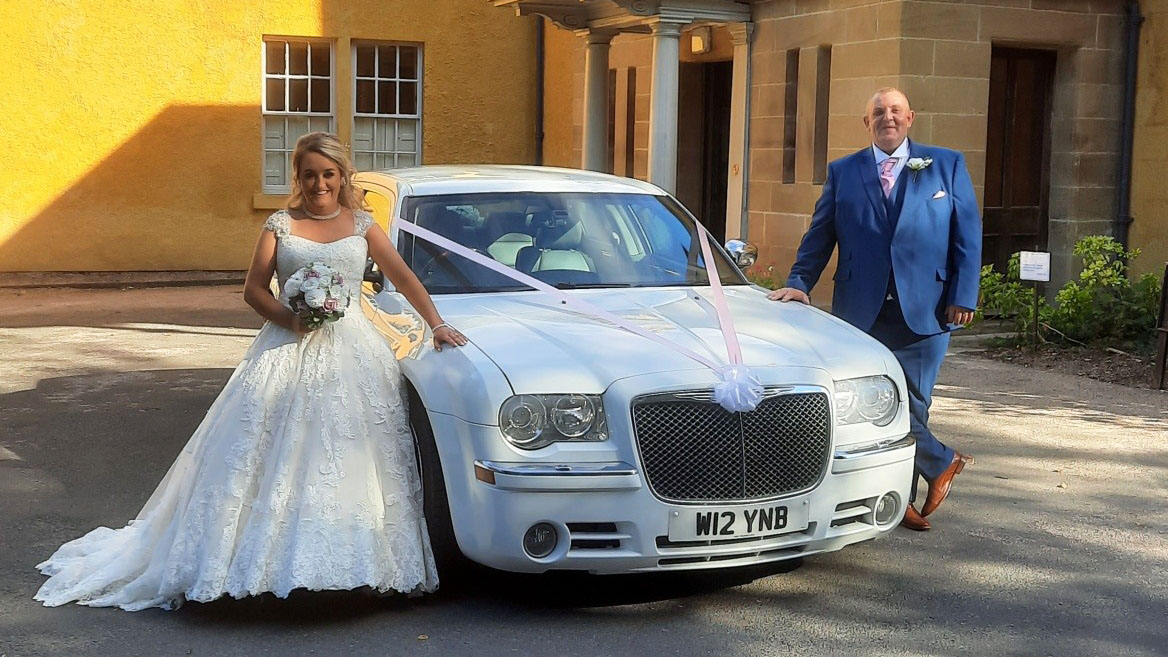 Bride and Groom standing on either side of a White Chrysler 300c Saloon Car decorated with white ribbons