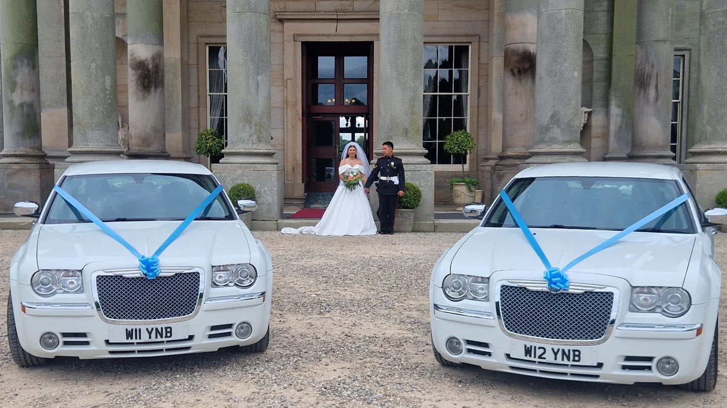 Two identical White Chrysler 300c Saloon Cars decorated with matching blue ribbons in front of a wedding venue with Bride and Groom in the background