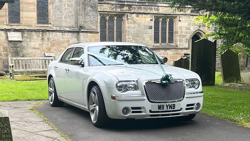 White Chrysler 300c Saloon Car with White and Royal Blue ribbons parked in front of a church in Durham