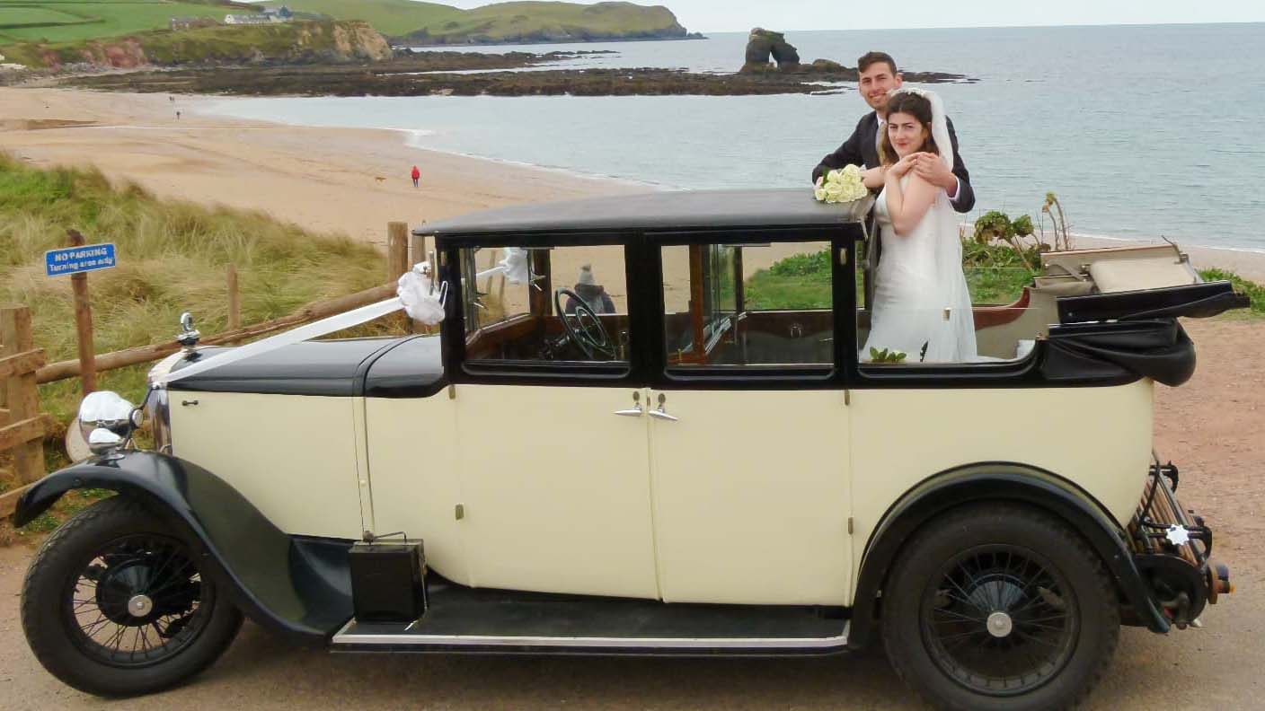 Vintage car with its convertible roof open parked on the Devon seaside with Bride and Groom standing inside the vehicle for photos