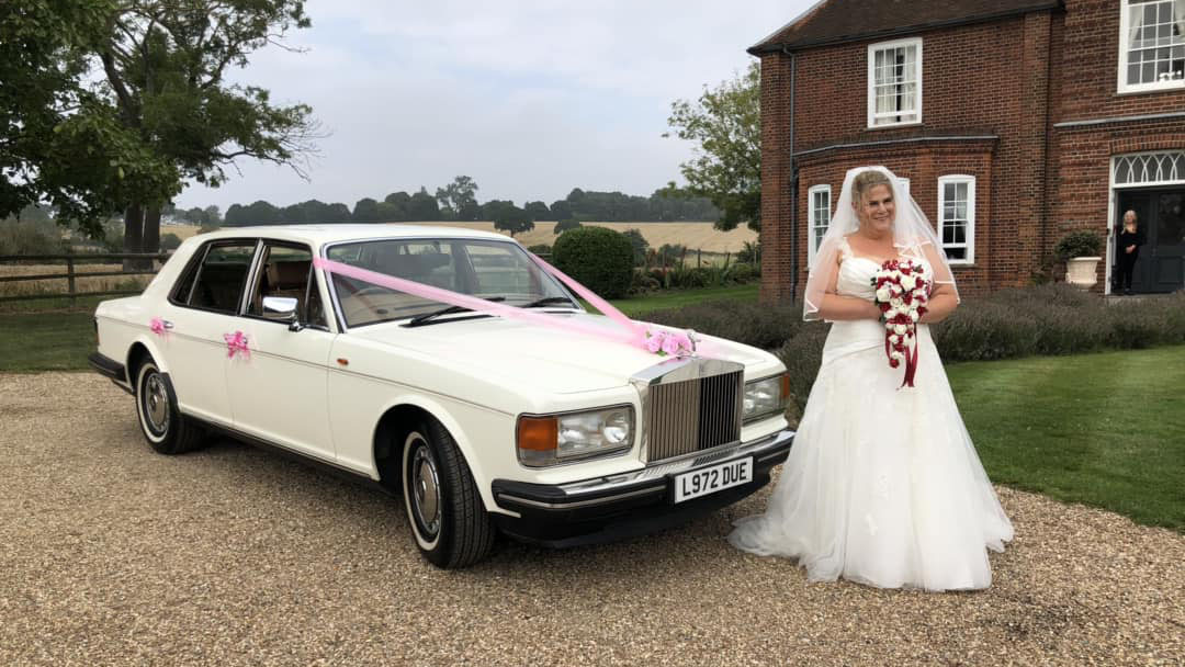 Bride wearing a white dress and holding a bouquet of white and red flowers standing in front of a classic Rolls-Royce Silver Spirit