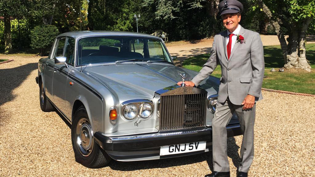 Chauffeur wearing a full uniform and hat standing in front of a classic silver Rolls-Royce Silver Shadow