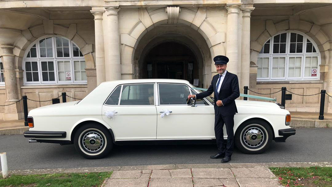 fully uniformed chauffeur wearing a hat standing in front of a classic Rolls-Royce Silver Spirit in white