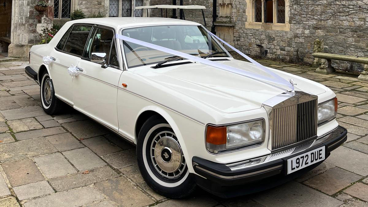 classic Rolls-Royce Silver Spirit in white dressed with light pink ribbons and bows parked in front of a wedding venue in Brentwood