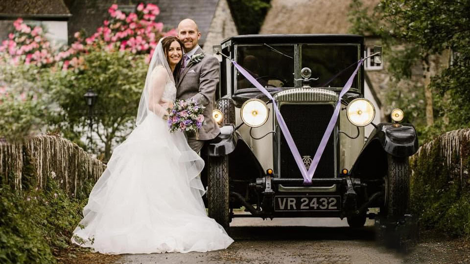 Bride and Groom posing for photos in front of a vintage daimler wedding car decorated with light purple ribbons and front headlights switch on.