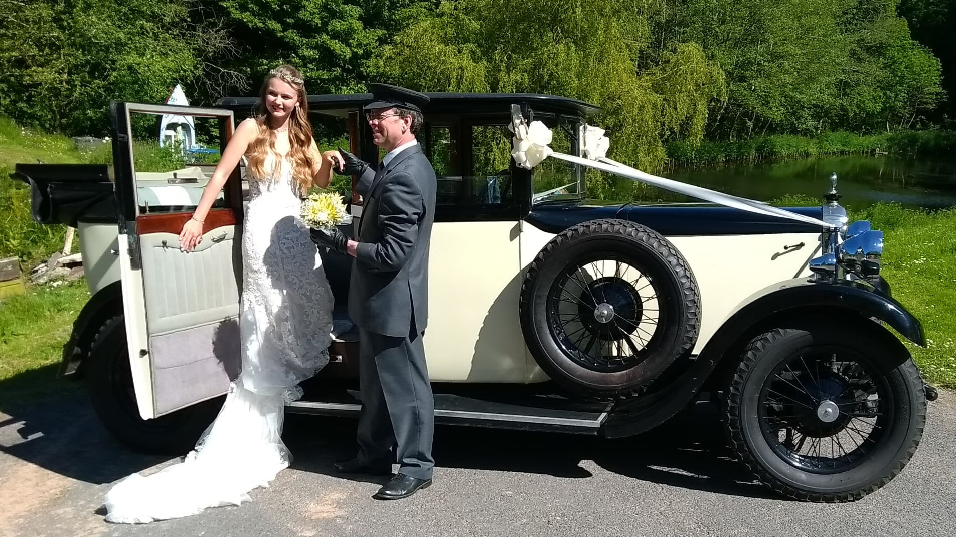 Bride wearing a white wedding dress exiting a black and cream car with the help of the chauffeur wearing a full uniform and hat.