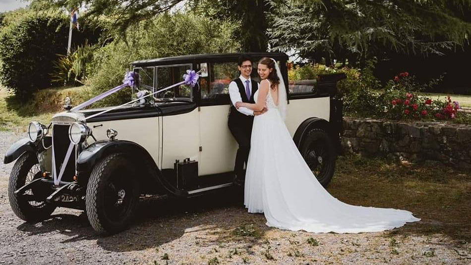 Bride and Groom holding each others in front of a vintage Black & Cream wedding car decorated with ribbons