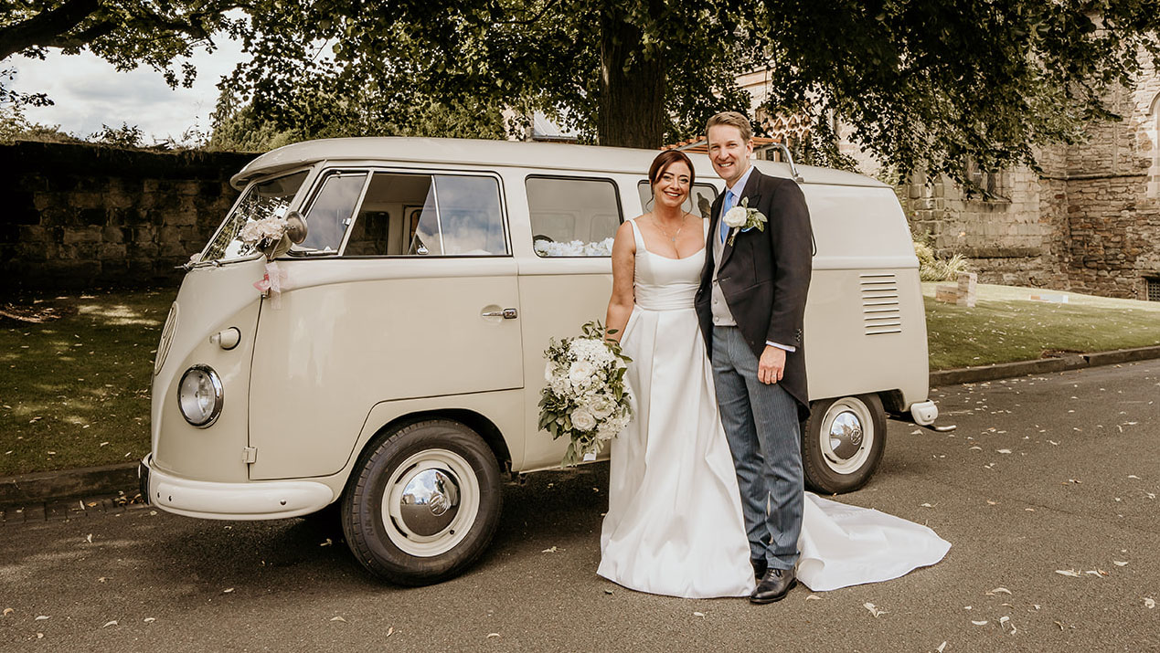 Bride holding a large bouquet of white flowers standing in frotn of a classic campervan with her groom