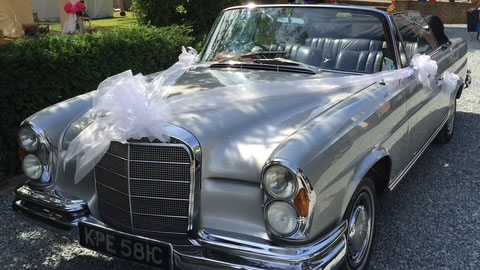 front left view of a classic silver Mercedes se220 convertible with white ribbons and large bow