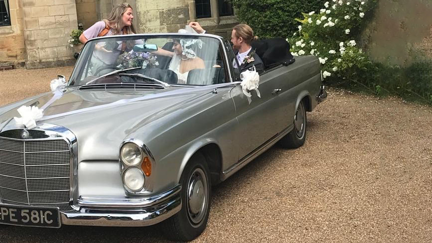 Bride and Groom seating in the rear of a classic convertible silver Mercedes decorated with white wedding ribbons and bows