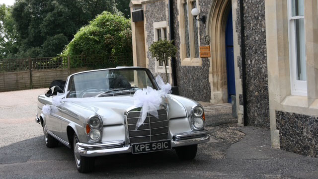 Silver Mercedes Cabriolet with a large white bow on top of its front radiator parked in front of wedding venue near Worthing