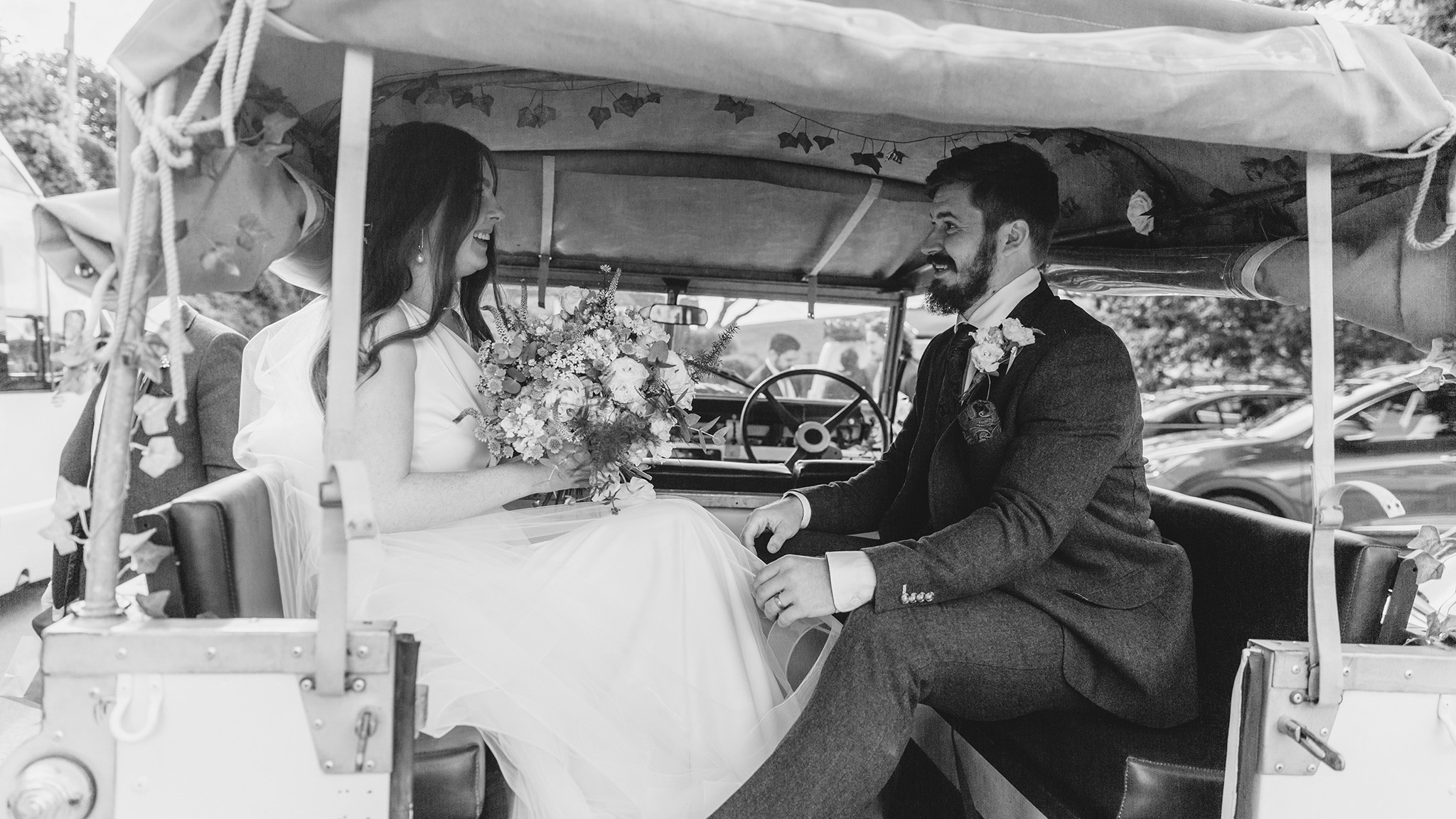 Black and White photo of Bride and groom seating inside a frotn view of Classic Land Rover with white ribbons and bows