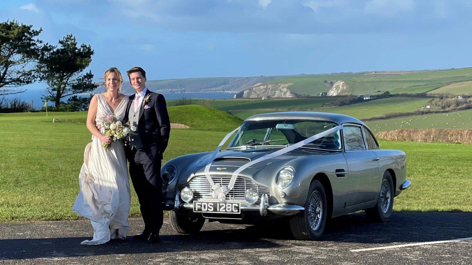 Bride and Groom standing in front of a classic Aston Martin dressed with white ribbons with Devon's sea coast in the background