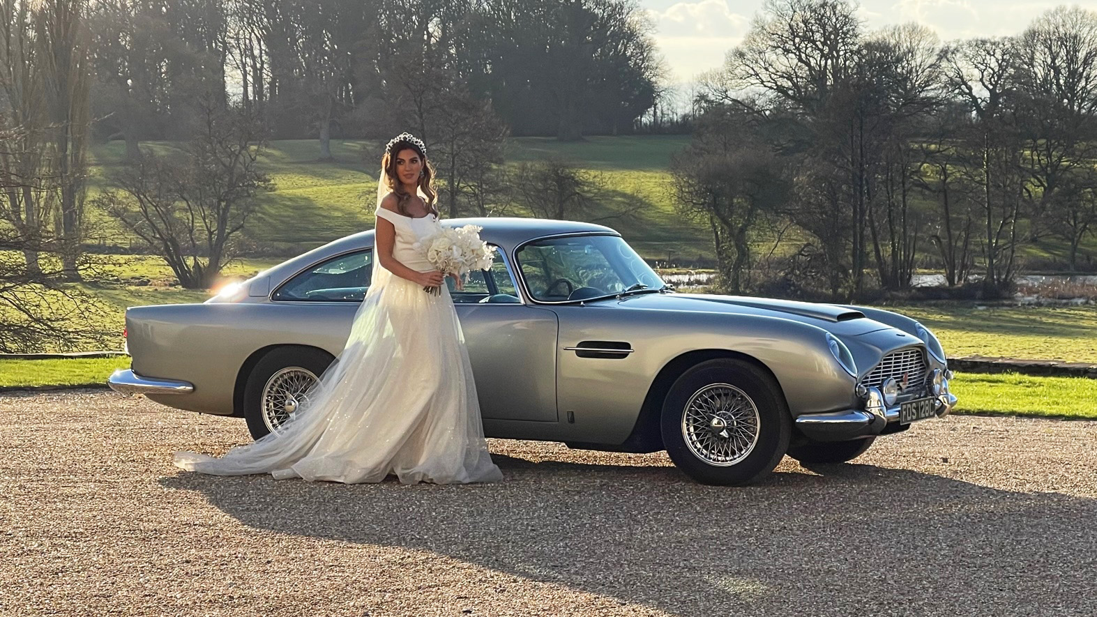 Bride wearin a white dress and holding a bouquet of flowers standing in front of a classic Aston Martin DB5