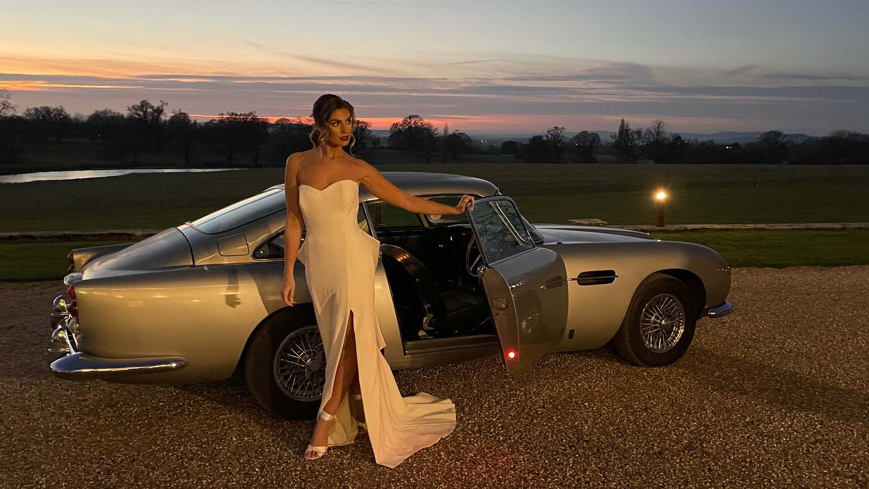 Bride wearing a white dress exiting a classic Astion Martin DB5 with a twilight background