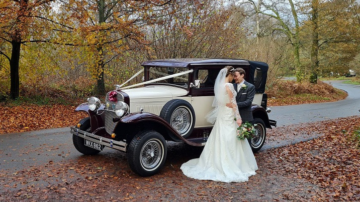 Bride and Groom kissing in front of a vintage Badsworth Convertible with White ribbons in an autumn forest background