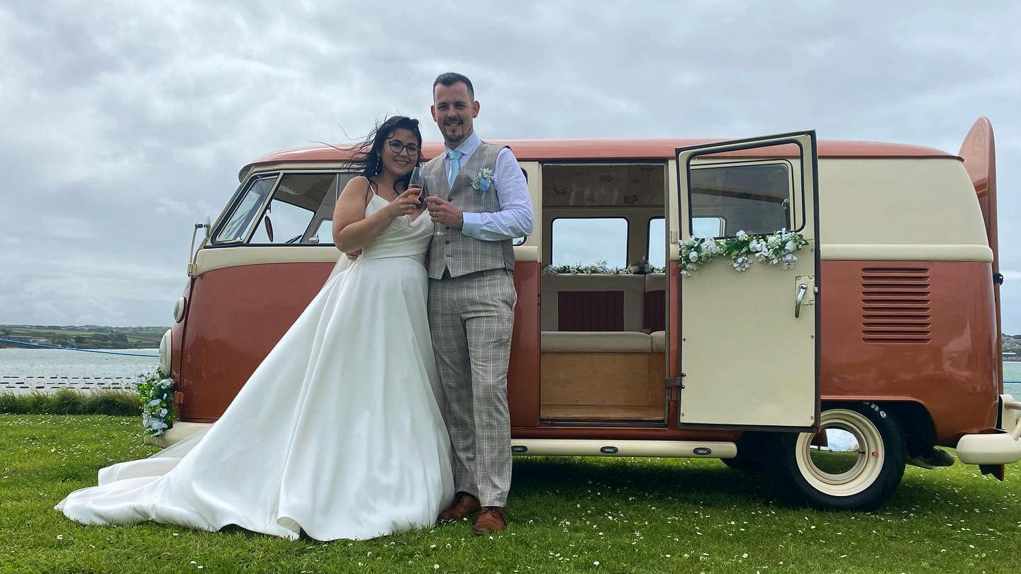 Bride and Groom standing in front of a classic VW Campervan in two tone Orange and white with rear passenger door open