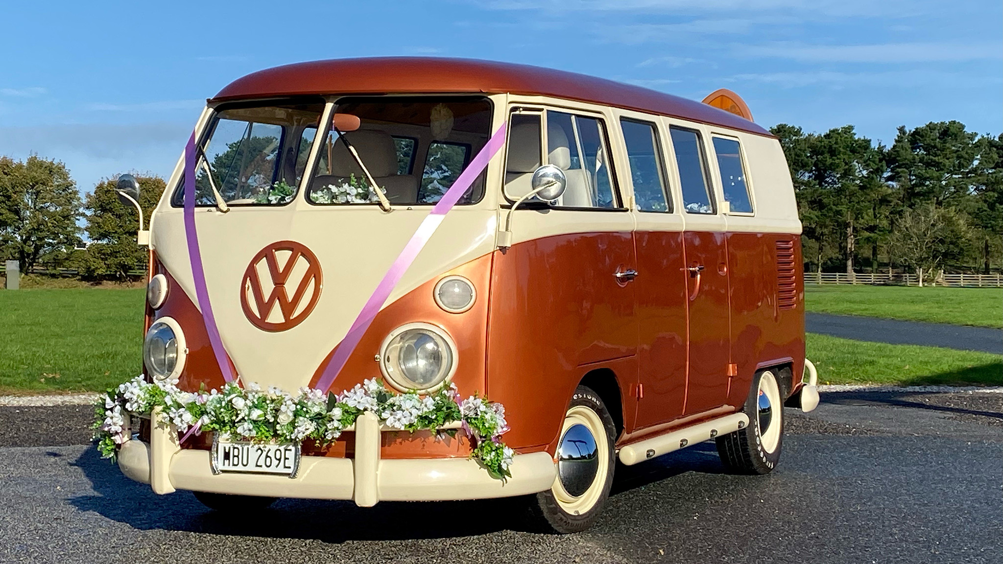 Classic VW Splitscreen campervan in two tone White and burnt orange decorated with green foliage on front bumper and white wedding ribbons